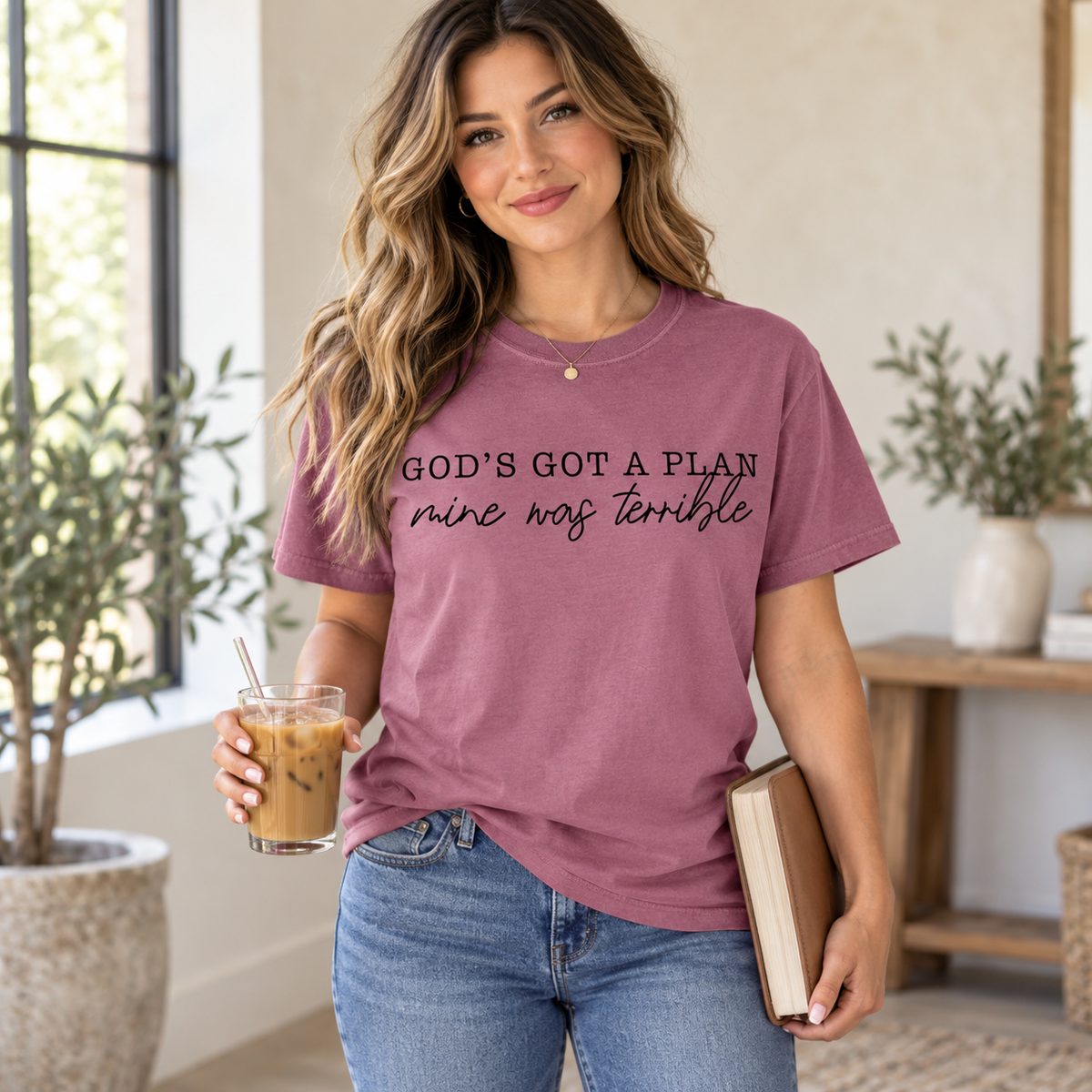 Standing woman wearing a mauve God's Got a Plan, Mine Was Terrible T-shirt, holding a closed Bible under one arm and iced coffee in her other hand.
