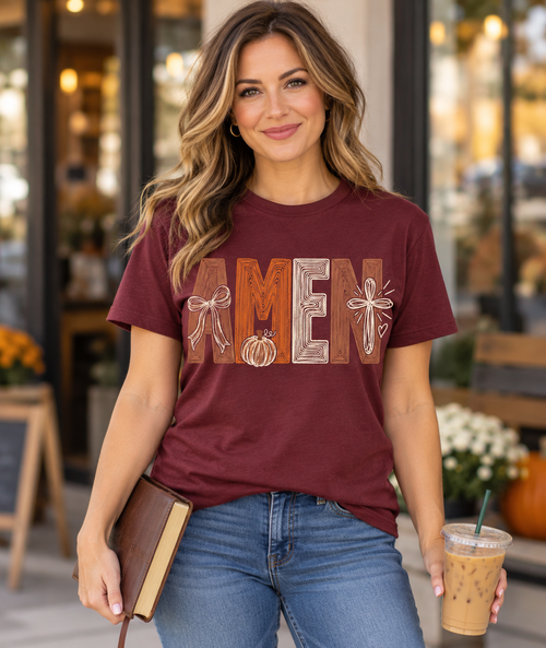 Model wearing maroon AMEN T-shirt with bow, pumpkin, and cross graphic, holding a Bible and iced coffee